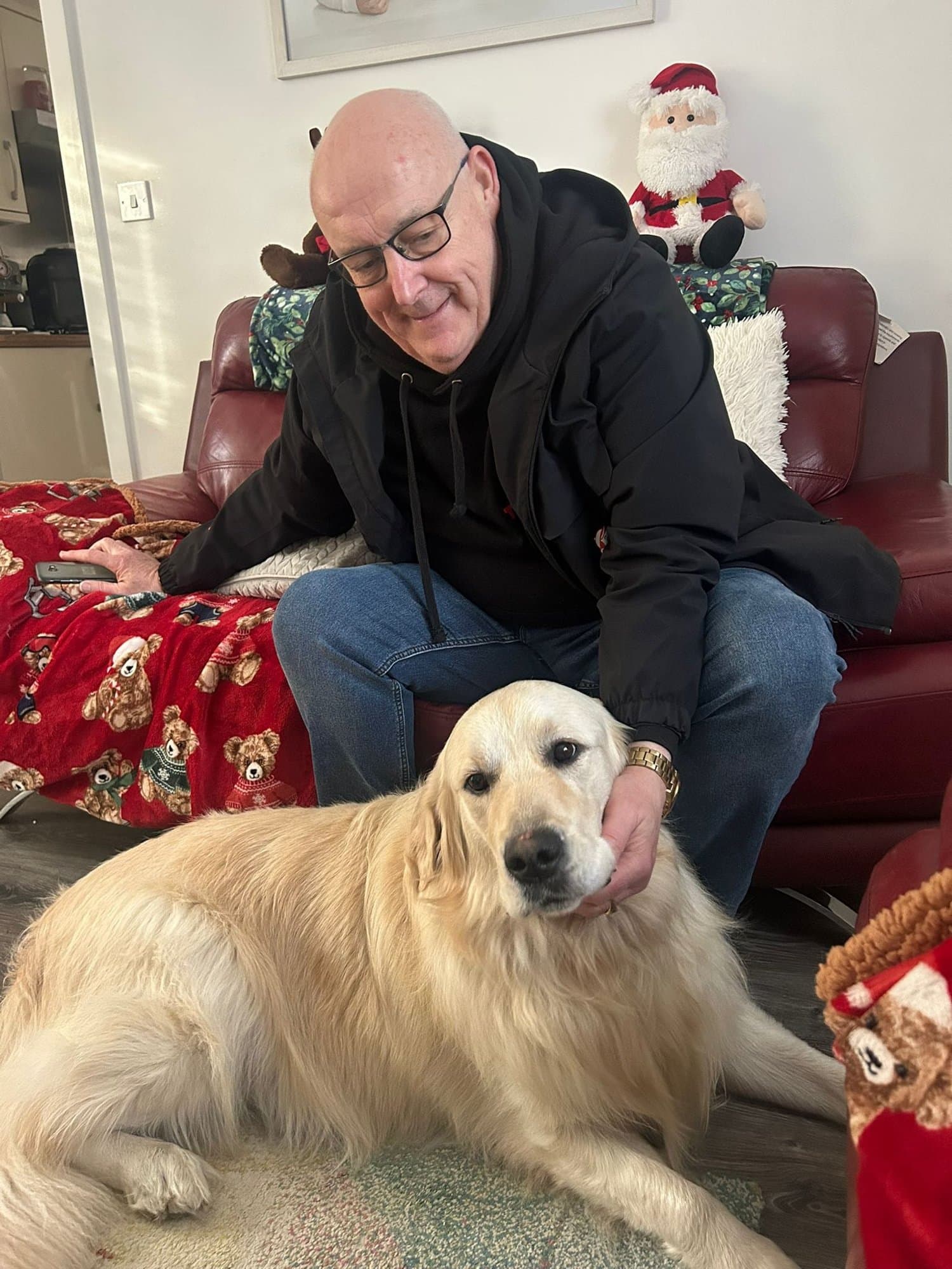 Brian smiling on a red leather sofa with his hand resting on a golden retriever lying at his feet